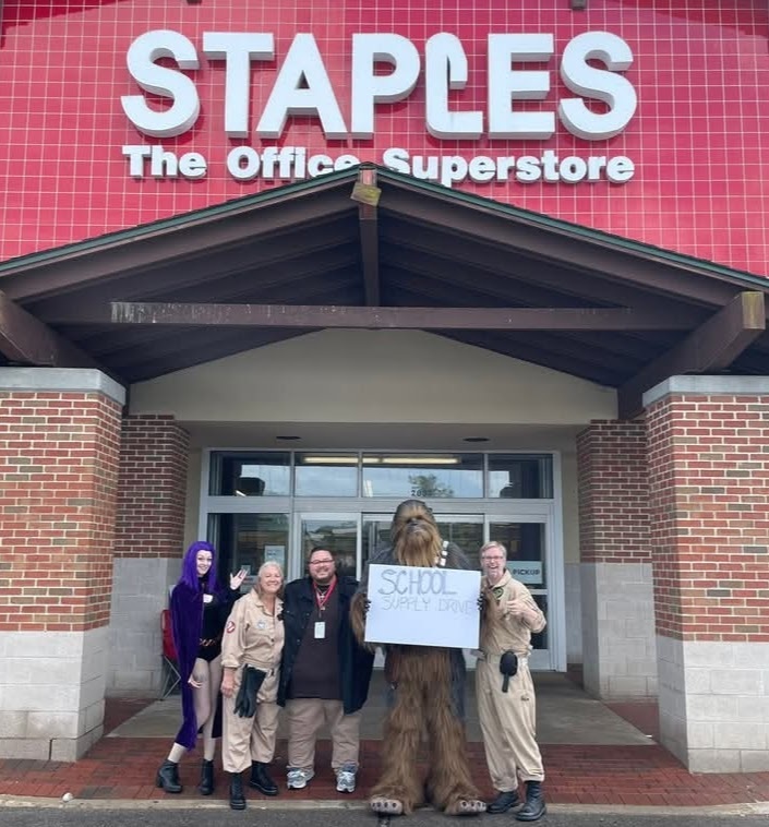 A group of five people, four in "Ghostbusters" uniforms and one in a Chewbacca costume, stand in front of a Staples store. The Chewbacca character is holding a sign that says "School Supply Drive."