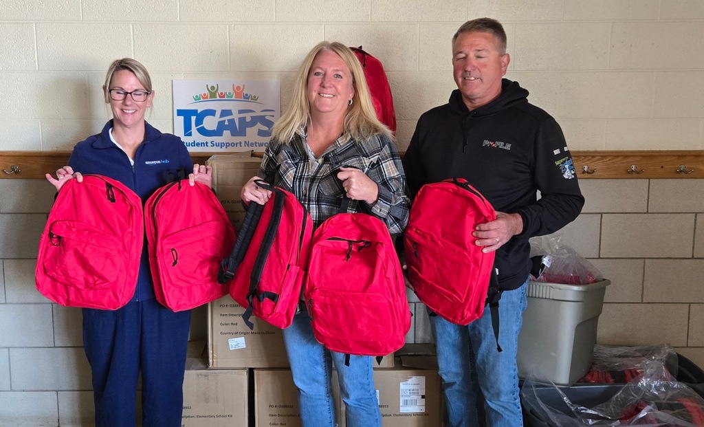 Three adults hold red backpacks in front of boxes and a TCAPS sign, representing a school supply donation.
