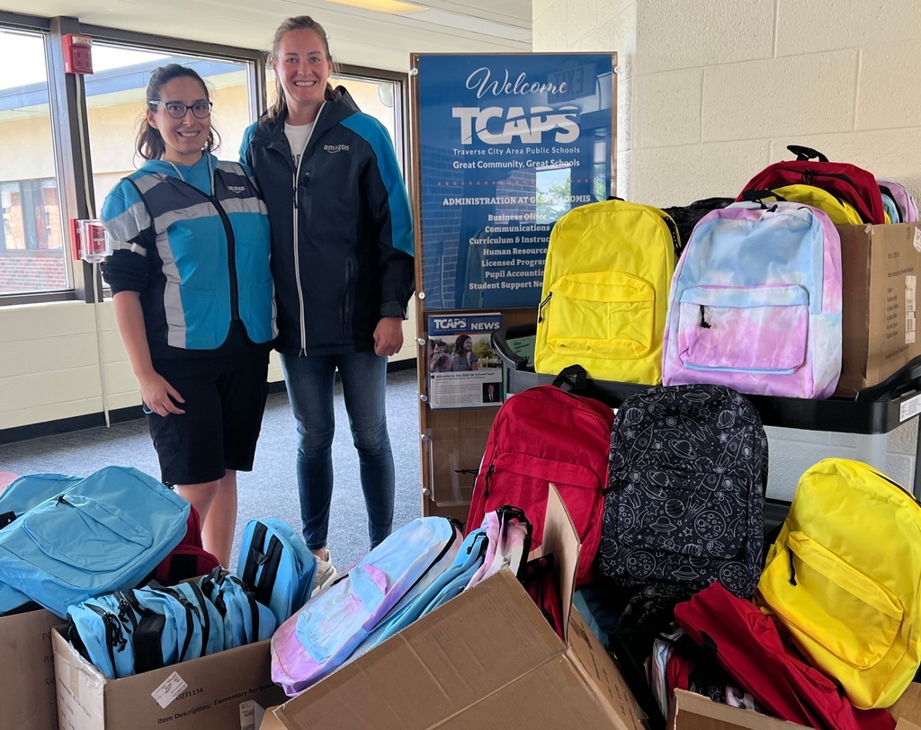 A photo of two women in Amazon jackets standing with a large donation of colorful backpacks for students.