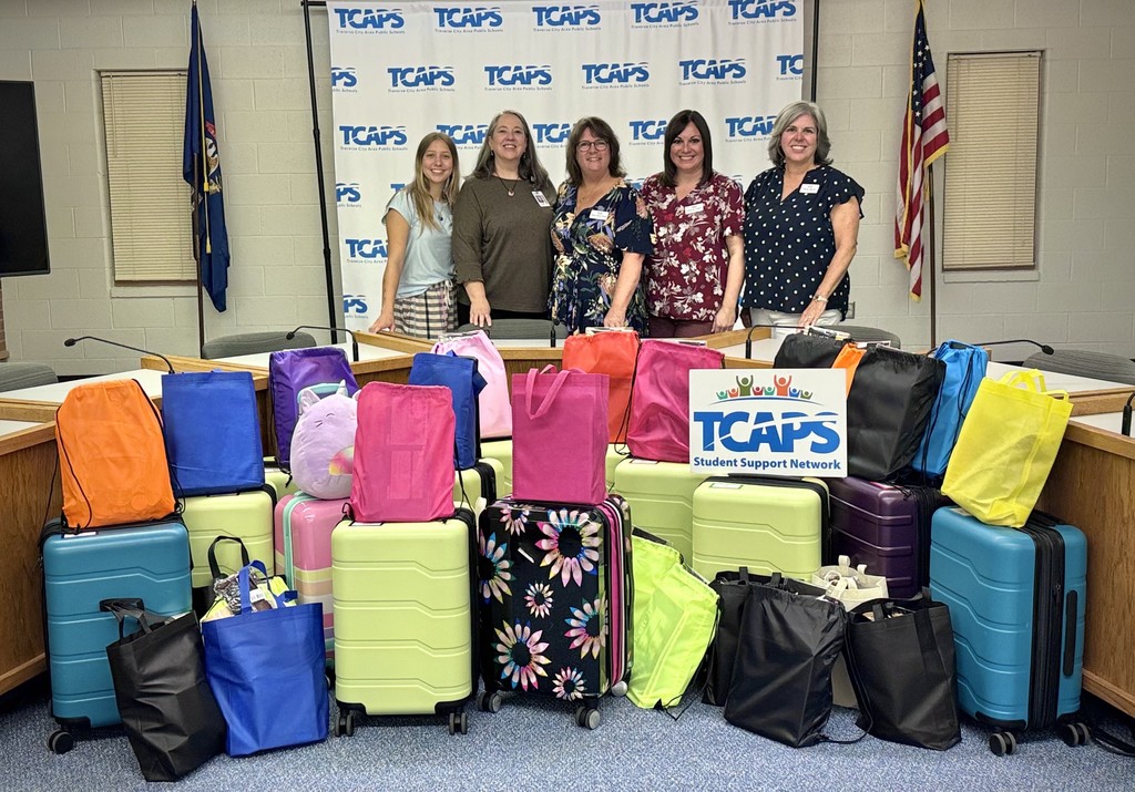Five women stand smiling behind a large donation of suitcases and bags for students.