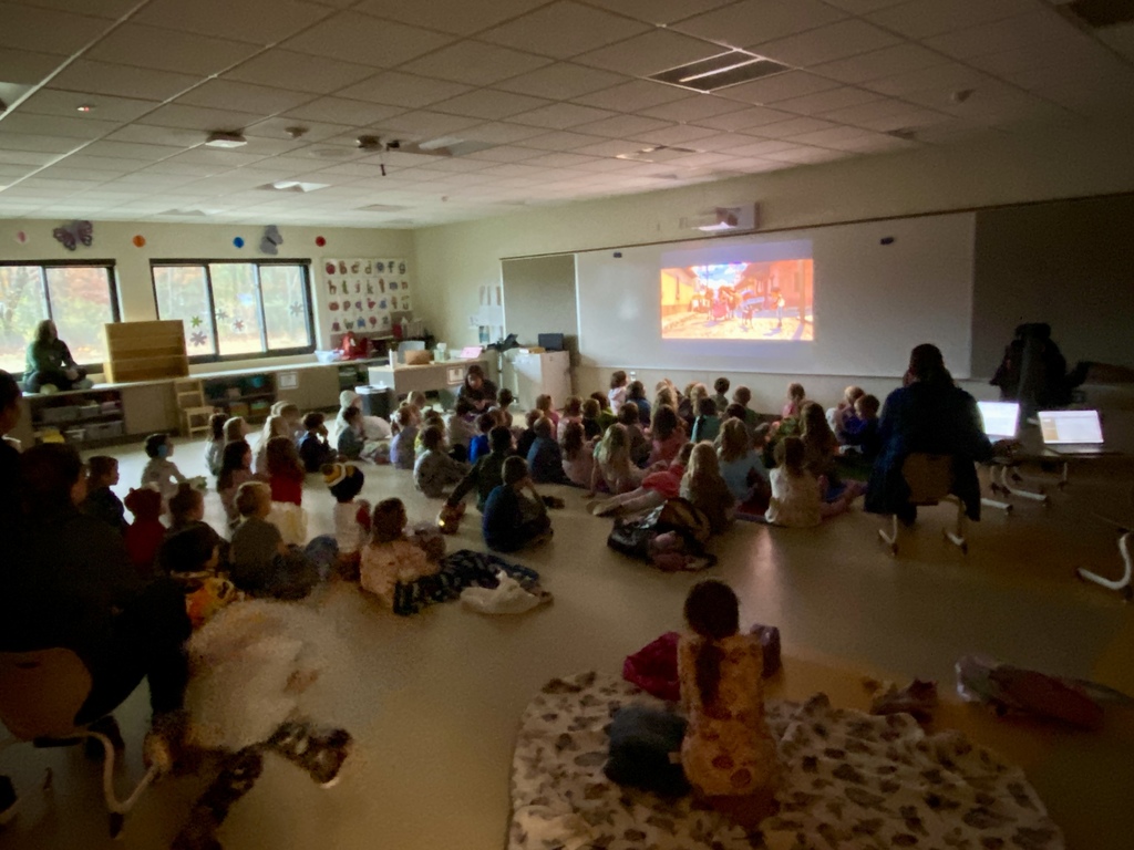 Students sitting on the floor in a classroom watching a movie projected on the whiteboard.  Teachers sitting in chairs.