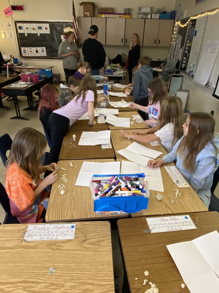 Students drawing animal pictures sitting at desks that are pushed together.  One student if reaching for some markers in a bin, while three adults in the back talk.