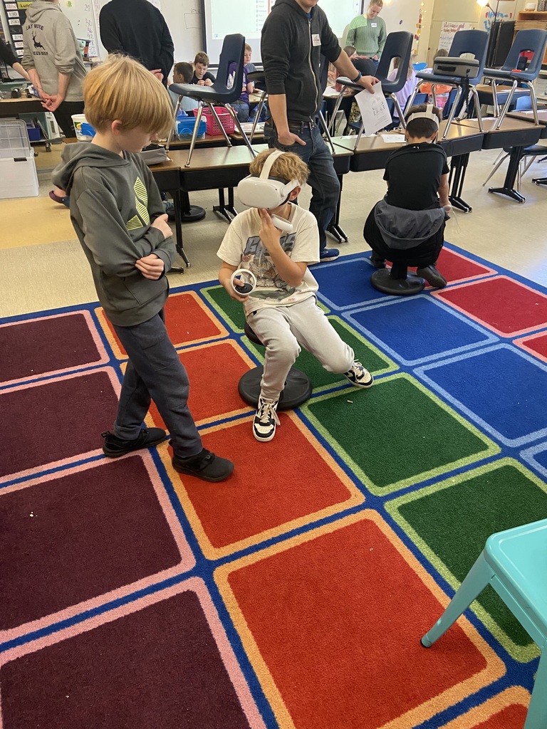 A student sitting on a black wobbly chair, which is on a colorful square rug, while another student looks on.