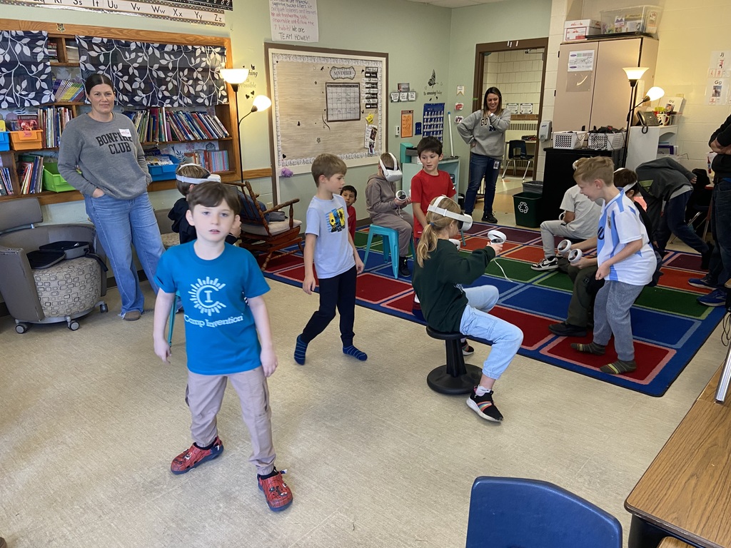 Students sitting on stools and wobbly  chairs using VR headsets, a few adults watching the students and one student looking at the camera.