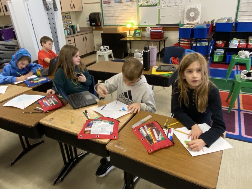 Students sitting at a desk with markers and paper coloring.  One looking at the camera and one student has a computer on their desk. 