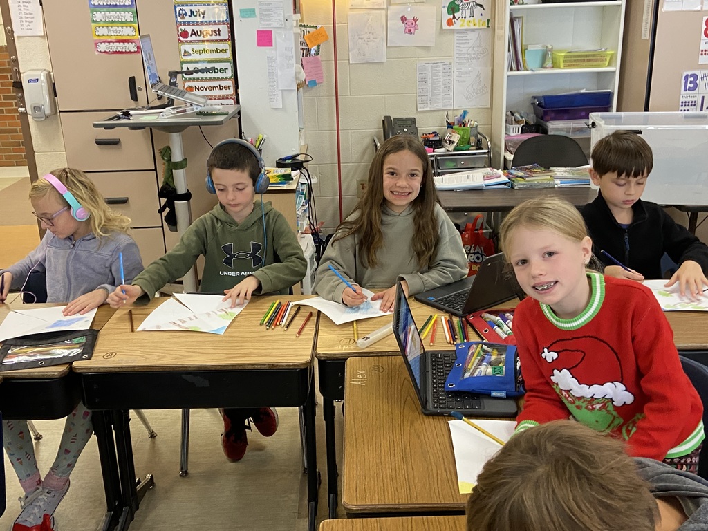 students sitting at their desk , coloring.  One student is wearing headphones and two are looking at the camera smiling