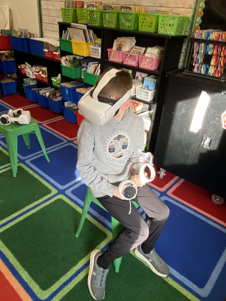 Student sitting on a green stool, on a colorful square rug, using a VR headset.