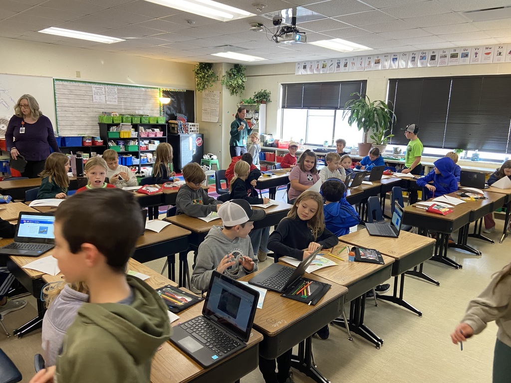 Students sitting at their desks, coloring and looking on their computer.  The teacher standing in the back helping another student. 