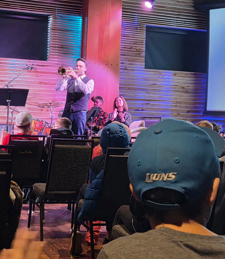 Students watching a guy playing the trumpet with a lady clapping behind him. 