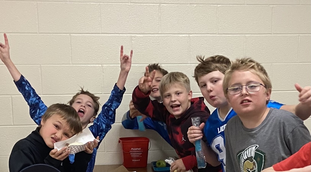 students smiling at the camera, some raising their hands, sitting in the lunchroom