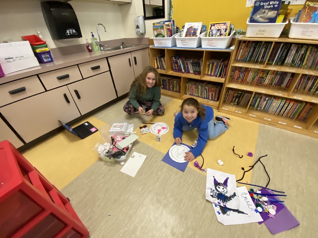 Two students sitting on the floor, in front of a bookshelf, make a craft on the floor smiling at the camera
