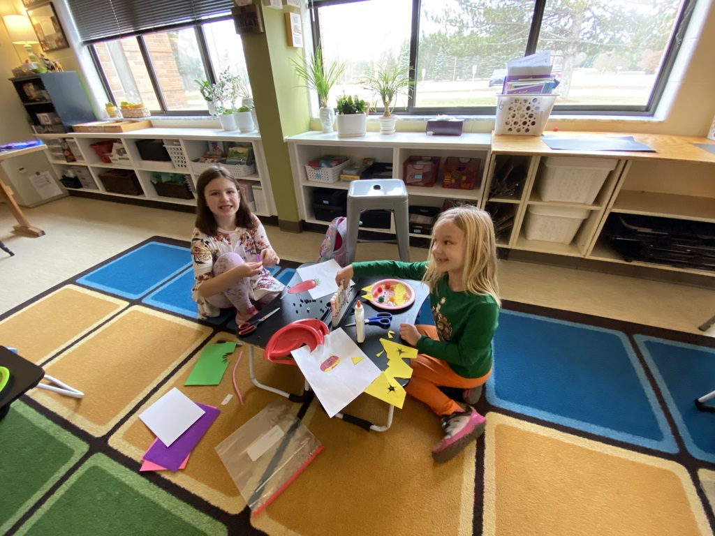 two students sitting on a square rug, the square rug has multiple squares of brown, blue, green and tan. The students are using a lapdesk on the floor, crafting. the students are smiling at the camera