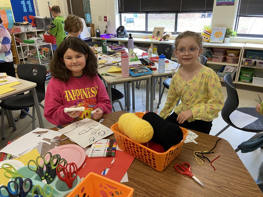 two students sitting at a table , a basket of yarn in between them, markers and glue. The girls are crafting.