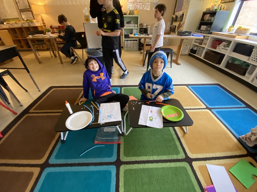 two students sitting on a square rug, the square rug has multiple squares of brown, blue, green and tan. The students are using a lapdesk on the floor, crafting. the students are smiling at the camera