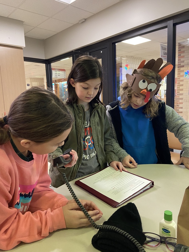 three students standing at the edge of the , reading a script while holding a phone, using the phone as a microphone to announce, one student wearing a turkey hat
