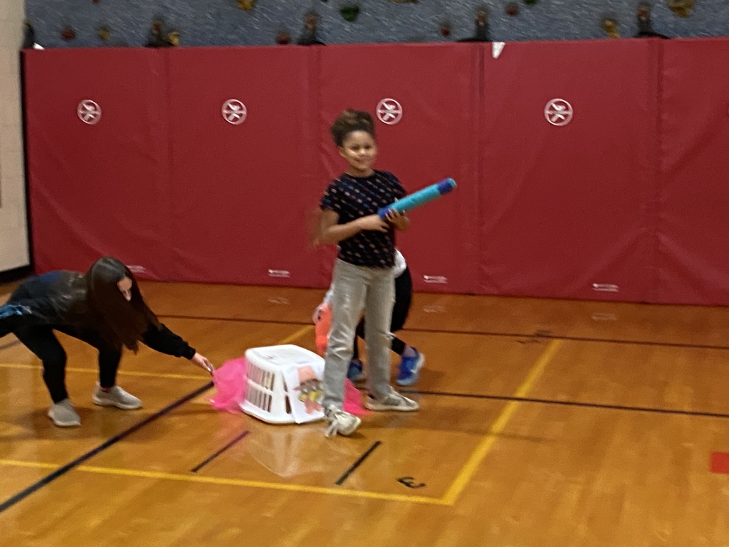 student standing with a pool noodle by a basket with a Tukey print out on the front with scarfs coming out of the side of the basket., another student trying to take the scarf.