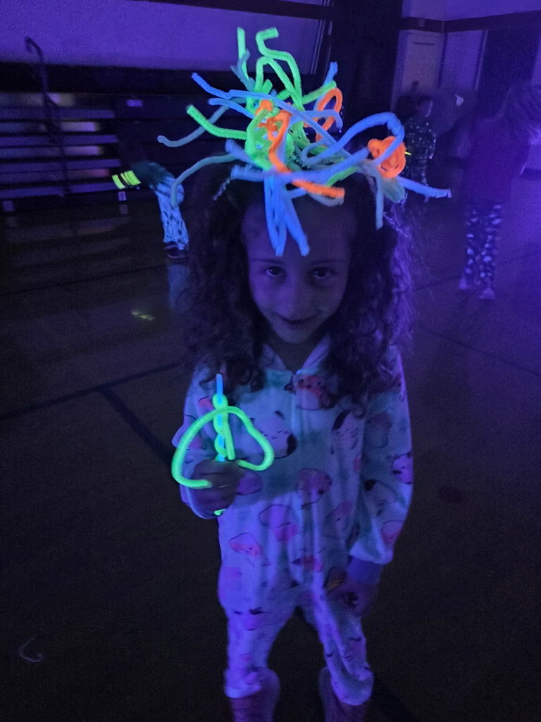 student wearing a "hat" of glow sticks on her head smiling for the camera in a dark gym for a glow party