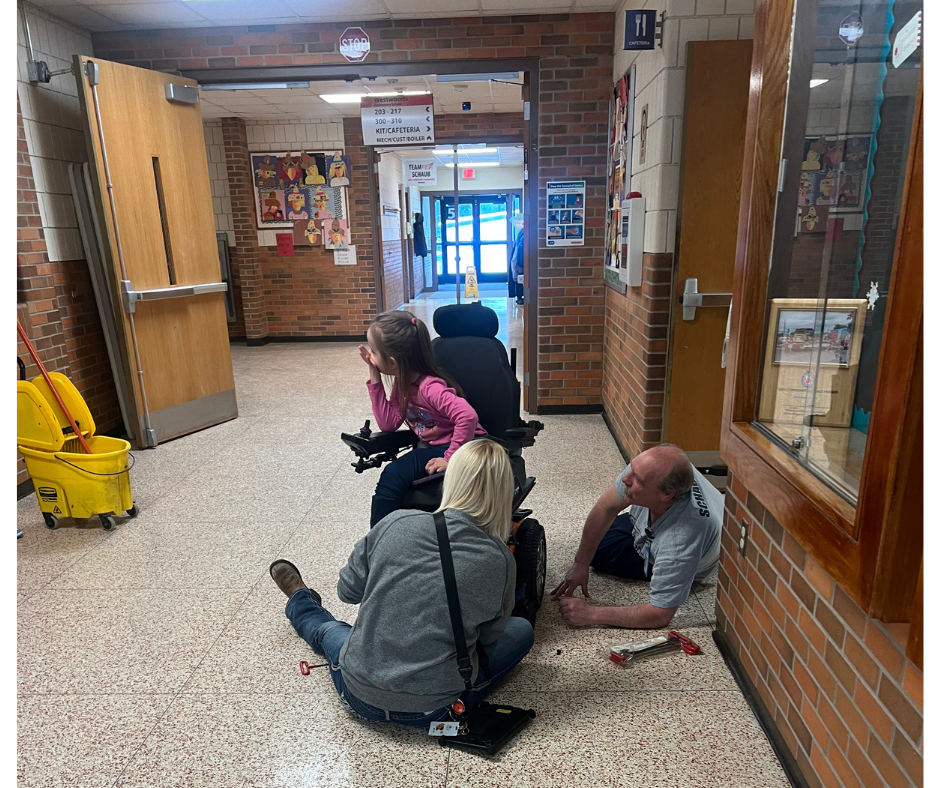 Mr. Sid laying on the floor helping to fix a wheel chair with Ms. Caity and a student in a wheelchair looking to the side.