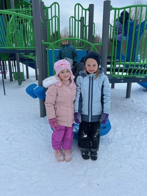 two students standing together outside in front the play structure smiling, one student behind smiling on the play structure. 