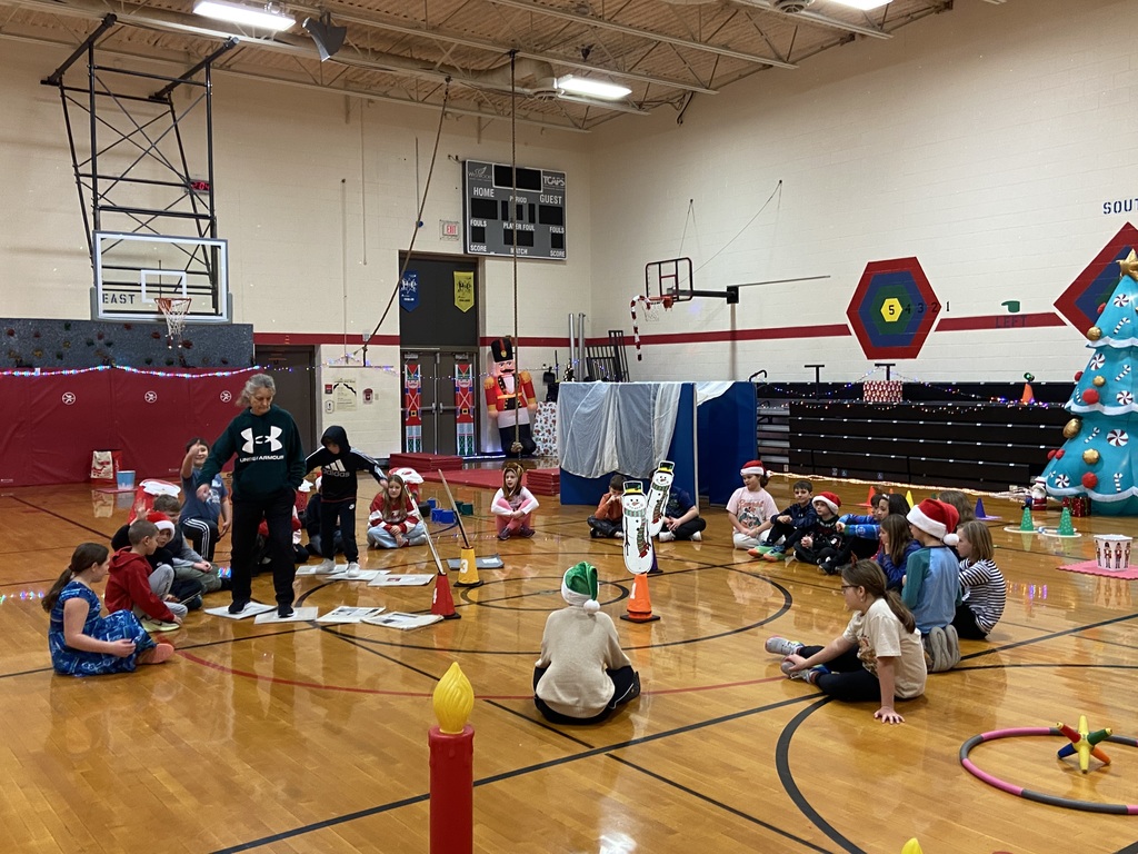students sitting in a circle at gym with Mrs. Ray teaching them.  The students are surrounded by a Christmas obstacle course.