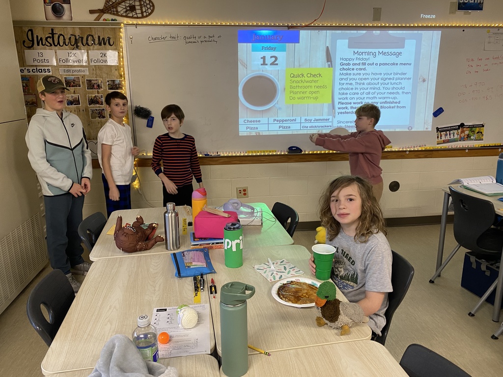 a student sitting at his desk eating a pancake and 4 other students in the background by the whiteboard.