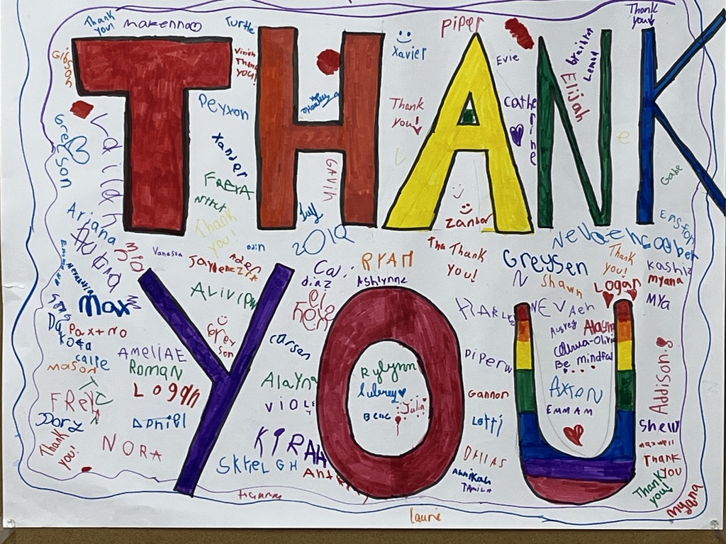 A close-up of a large, hand-drawn "THANK YOU" poster on white paper. The letters "THANK YOU" are written in a variety of solid colors including red, orange, yellow, green, blue, and purple, with the final "U" featuring a rainbow design.