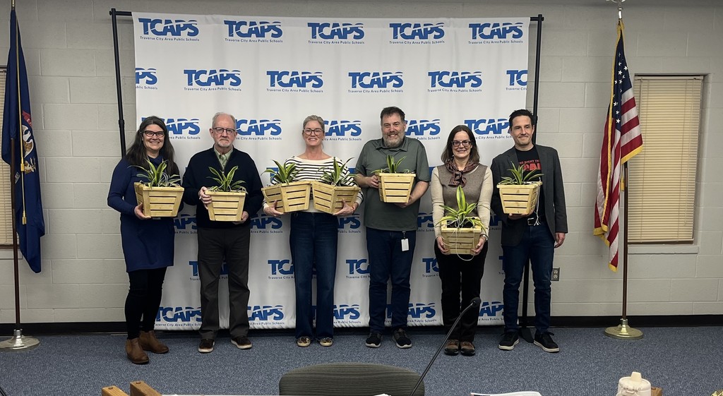 The six TCAPS Board of Education members are posed in a wide shot in front of the TCAPS logo backdrop. In this photo, each member is smiling and holding a light wood planter box containing a green leafy plant. 