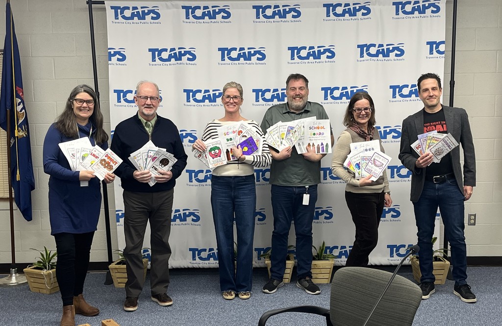 A group of six TCAPS Board of Education members stand in a row against a white backdrop featuring the repeating TCAPS logo. Each member is smiling and holding a large stack of colorful, hand-drawn "Thank You" cards and letters created by students.