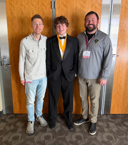 Three men standing indoors against a light wood-paneled wall. Porter Lawens in the center wears a black tuxedo and a gold medal on a yellow ribbon. To his left is Doug Downer, and to his right is a man with Chad Mielens.
