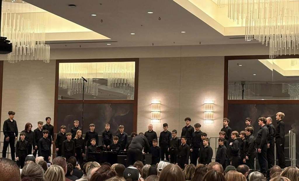 A student tenor and bass choir performing on a stage for an audience in a hall with modern chandeliers.