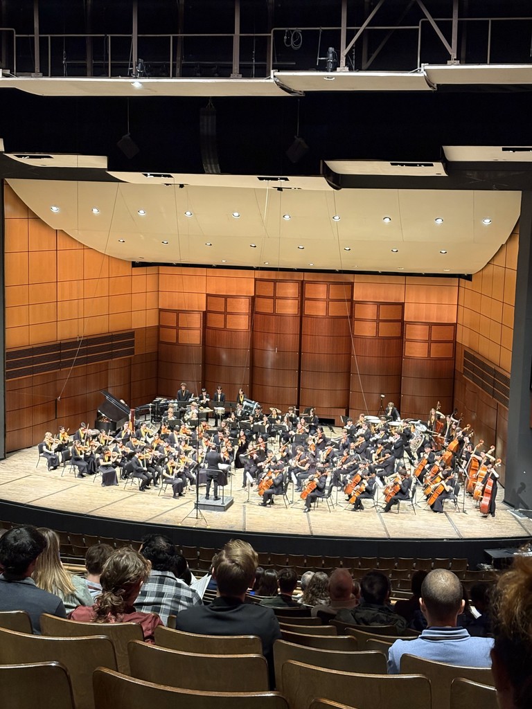 A student symphony band performing on a professional wood-paneled stage with a conductor at the front.