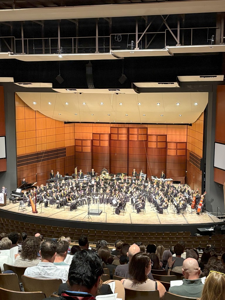 A student symphony band performing on a professional wood-paneled stage with a conductor at the front.