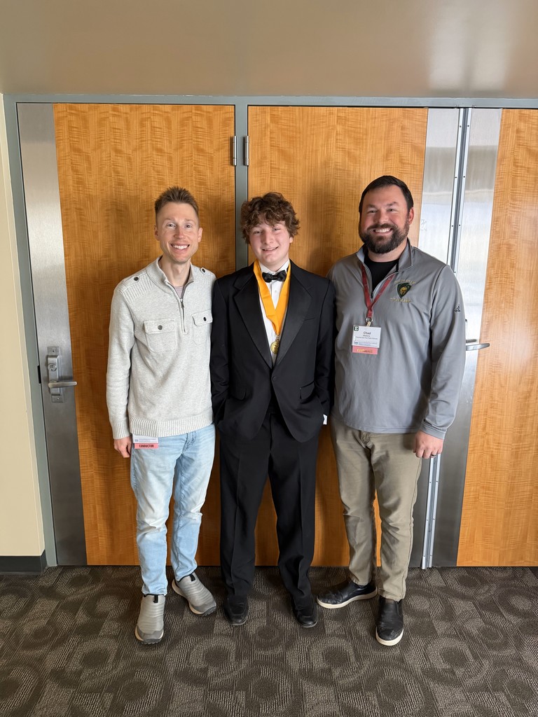 A student in a tuxedo and All-State medal standing between two smiling instructors in front of a wood-paneled door.