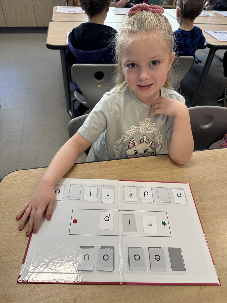 A young student smiling at the camera while sitting at a school desk. They are working on a CKLA activity with a white board containing various letters and phonics tiles.