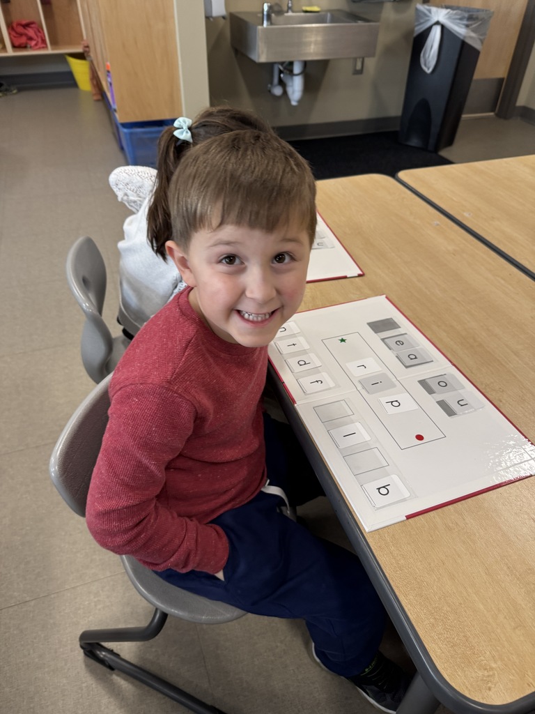 A young student smiling at the camera while sitting at a school desk. They are working on a CKLA activity with a white board containing various letters and phonics tiles.