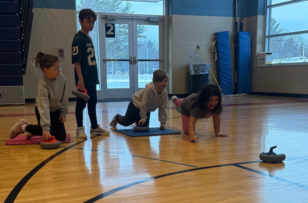 Four students in the gym, all holding curling rocks, one student has released the rock across the floor.