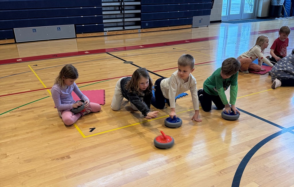 Another group of four students crouching on the gym floor; three have curling rocks and one has released it from their hand.