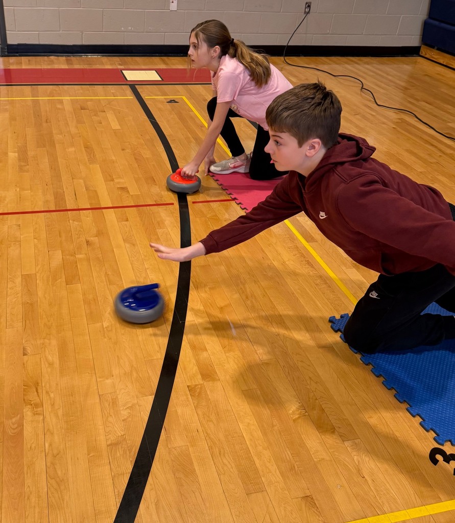 One female student and one male students crouched on the gym floor. The male student has released a curling rock from his hand and the female student is waiting to release her curling rock.