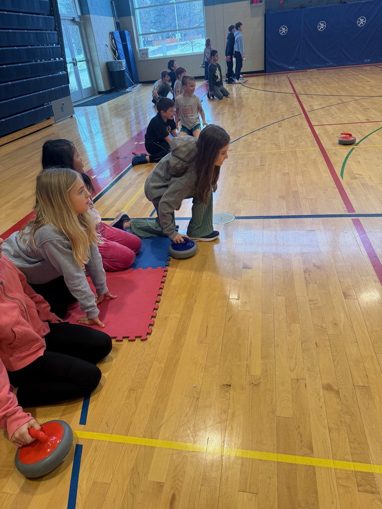 A group of elementary students in a line crouching on the gym floor, two have curling rocks in their hands waiting to release them to slide across the gym floor to a target not pictured.