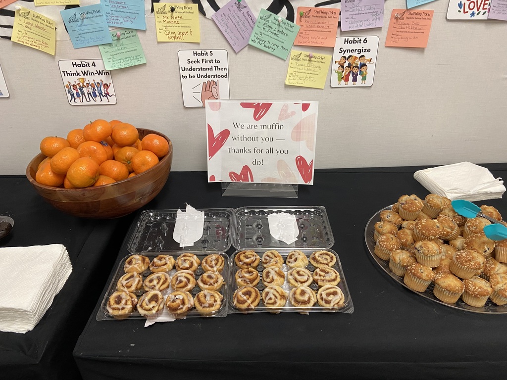 A oranges in a wodden bowl, a tray of cinnamon rolls and a plater of muffins on a black table cloth with a sign reading: We are Muffin without you thanks for all you do!