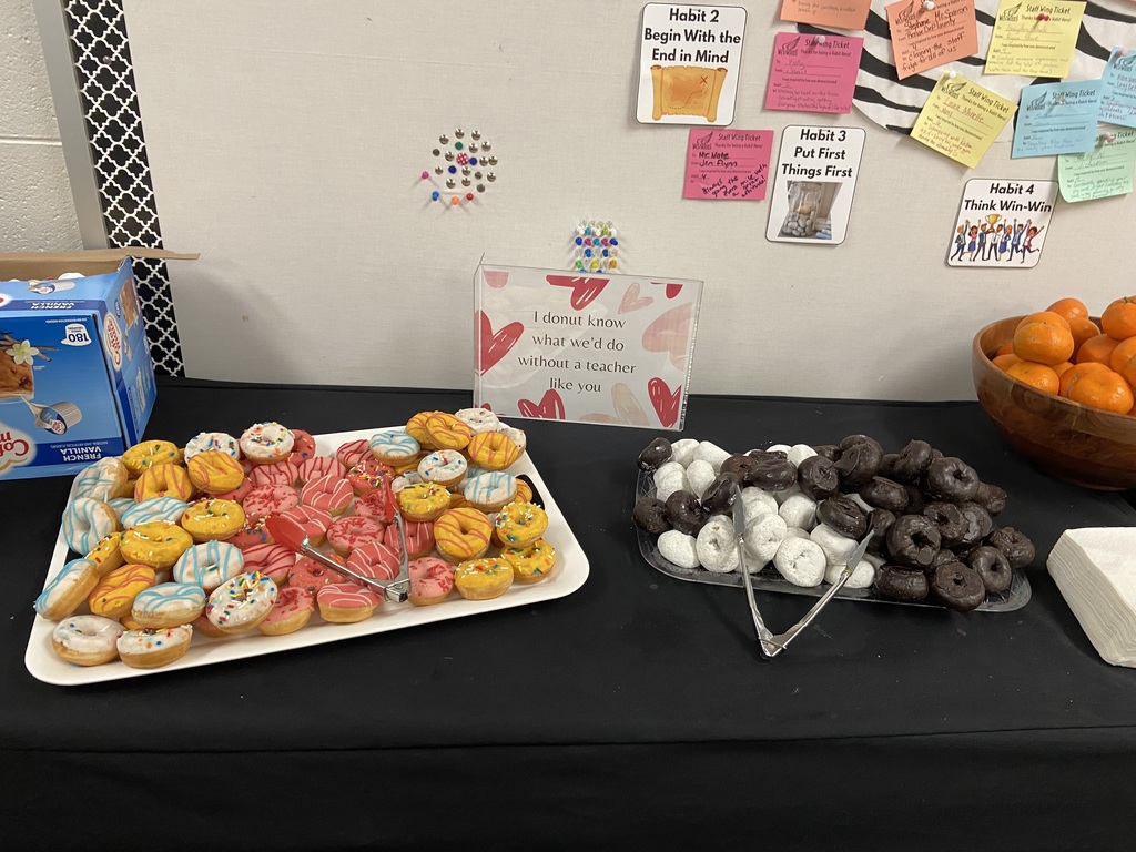  Two trays of donuts, one with white and black donuts, the other one with yellow, pink, ble and white donuts on a black table cloth, with tongs. A bowl of oranges on the side and a sign reading: I donout know what we'd do without a teacher like you!