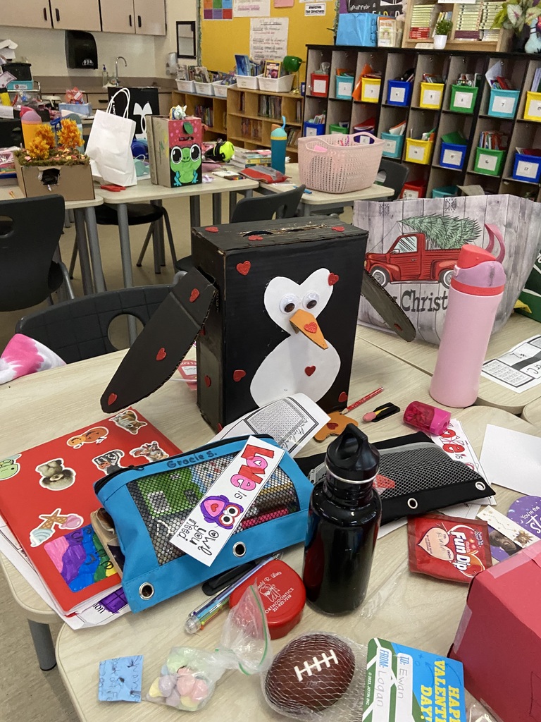 A box covered in black paint and a penguin on the front with two eyes, a orange nose, two wings and small red hearts spartically on the box.  The desk is covered with pencil bags, water bottles, red folder, a red retainer case, pencils and papers.