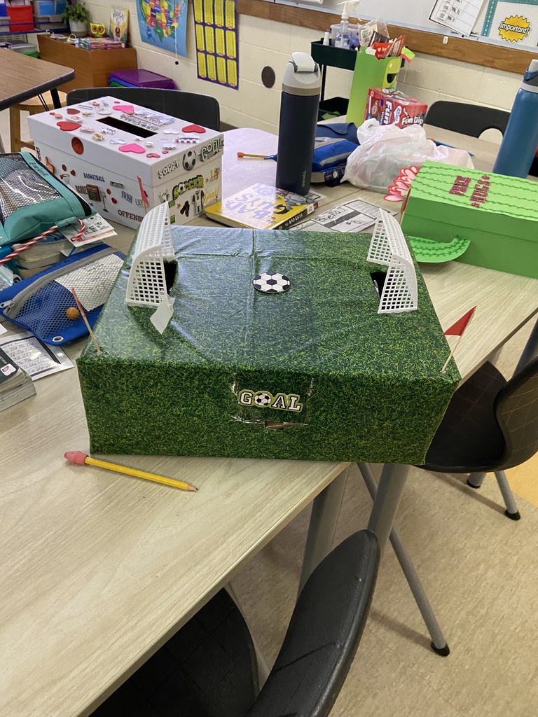 A box covered in paper that looks like grass with two soccer goals on the top, a soccer ball in the center and the word goal written on the side.  Red flags are sticking out the edge of the box.