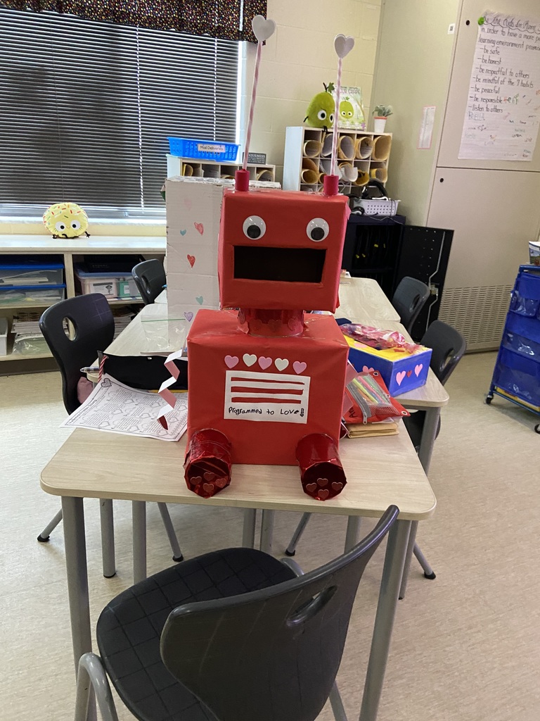 A red robot valentines box with white and pink hearts, two eyes, pink arms and red little legs that look like cans, with hearts on them.  The box has a white sign on the front and it reads programed to love with red stripes above it.  Behind the desk is tanother box that is blue and yellow with hearts on it and another box with hearts on it.  