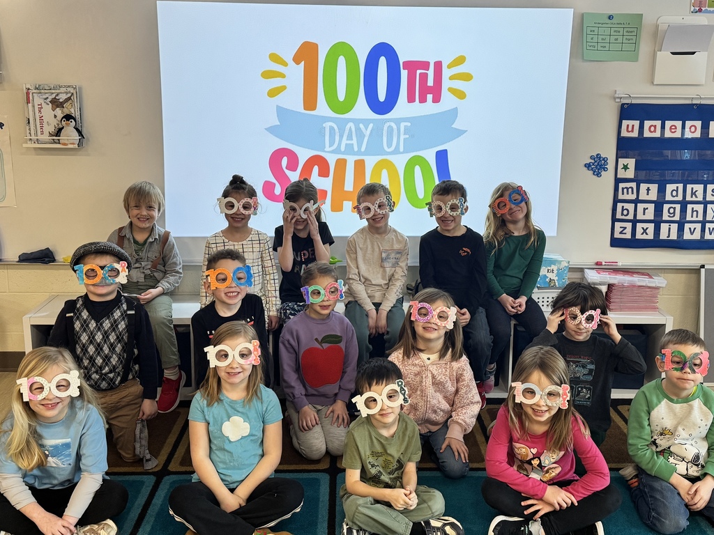 students lined up in front of 100th day of school projected on the whiteboard.  The students are sitting, smiling for the camera wearing 100th day of school sunglasses.
