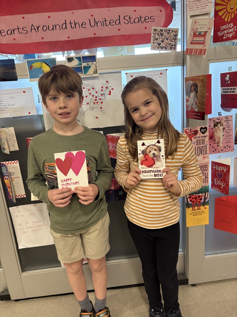 Two second grade students, one boy and one girl, each holding a Valentine card in front of their bodies with two hands.