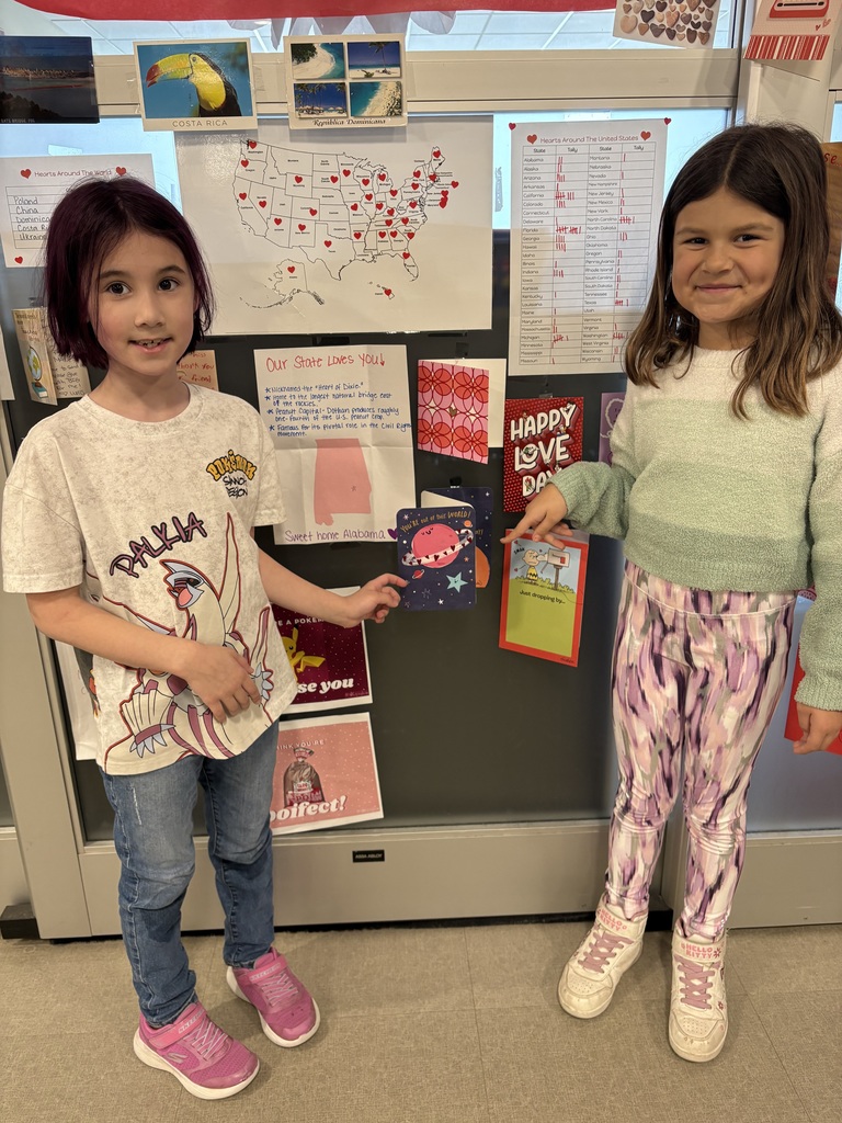 Two second grade girls standing in front of a map of the USA that has red dots indicating the 37 states from which they received valentines and also using their hands with a pointed finger towards the valentines.