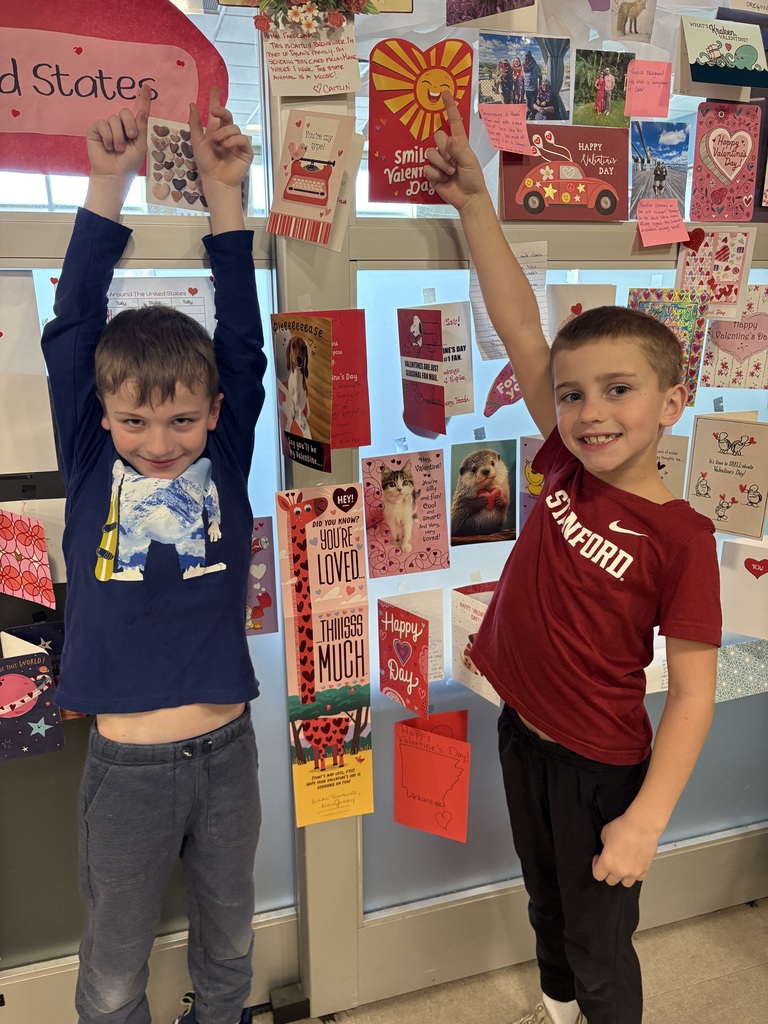 Two second grade boys standing in front of a window wall covered with Valentines taped to it. The boys are holding up their arms pointing to the valentines.