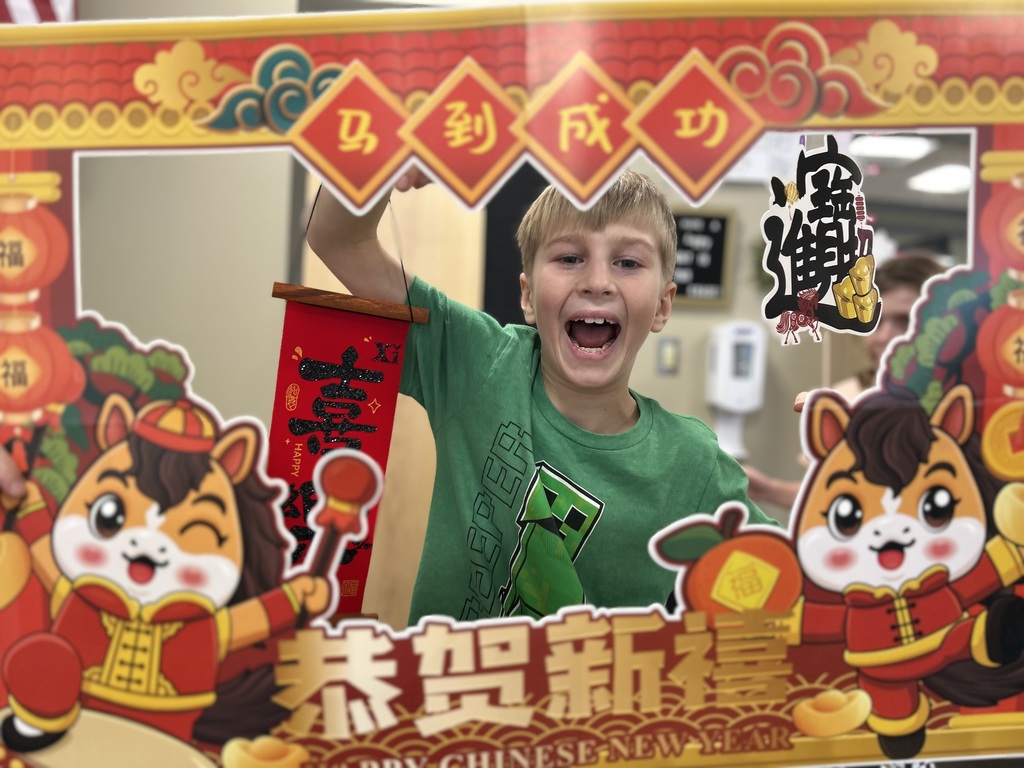A second grade boy looking through a festive cardboard frame decorated for the Chinese New Year and he is holding a small red banner with Chinese language symbols on it..