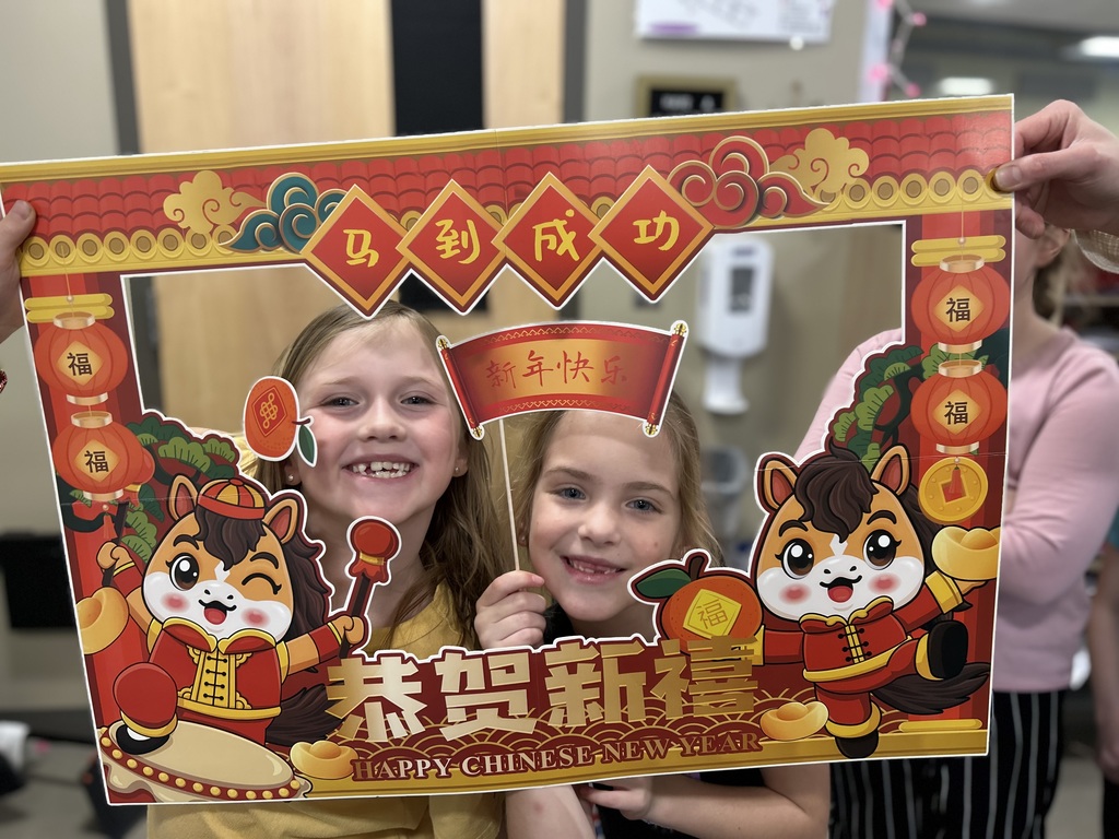 Two second grade girls looking through a festive cardboard frame decorated for the Chinese New Year that they are holding.